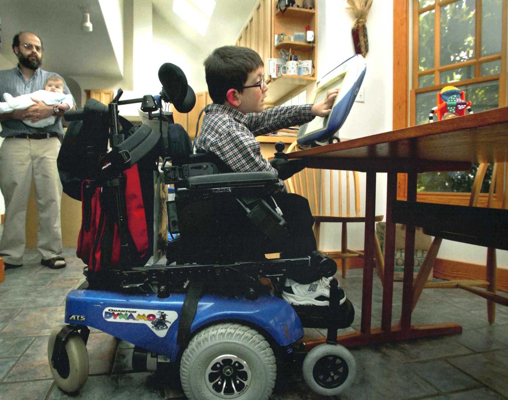 A young boy sits in a wheelchair manipulating a portable computer screen. A middle aged man holding a baby looks on.