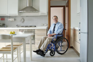 A businessman wearing a tie sits in a wheelchair in his kitchen at home.